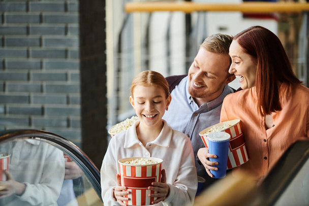 映画館のエスカレーターの隣に立っている間ポップコーンのバケツを握る家族. - 写真・画像
