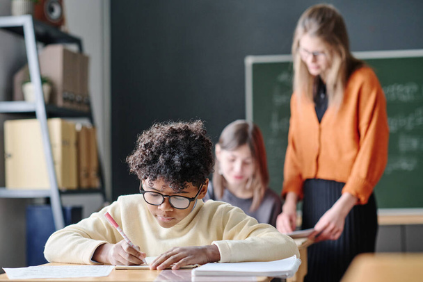 Selective focus shot of African American teen boy writing in notebook during lesson, young teacher helping girl with task in background - Photo, Image