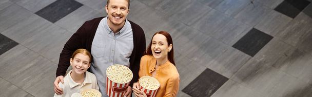 Mężczyzna, kobieta i dziewczyna szczęśliwie trzymają pudełka popcornu w kinie. - Zdjęcie, obraz