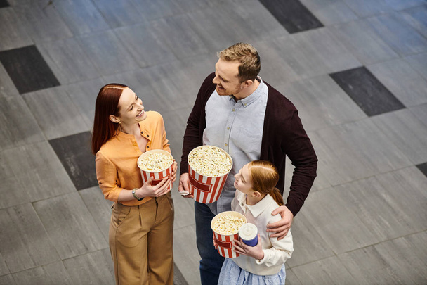 Ένας άντρας και μια γυναίκα κρατούν με ενθουσιασμό κουβάδες ποπ κορν ενώ ένα παιδί χαμογελά, δημιουργώντας μια ευτυχισμένη οικογενειακή σκηνή στον κινηματογράφο.. - Φωτογραφία, εικόνα