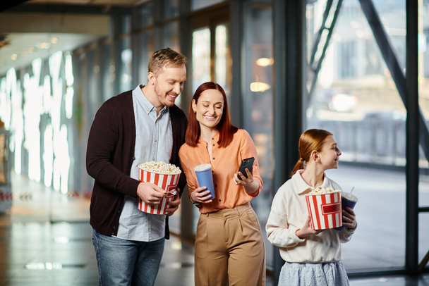 Ein Paar hält fröhlich einen Eimer Popcorn in der Nähe seiner Tochter und genießt die gemeinsame Zeit im Kino. - Foto, Bild