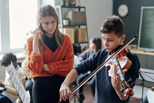 Young Caucasian female teacher listening to her student playing violin during music class at school - 写真・画像