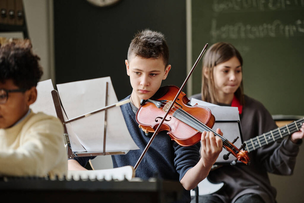 Selective focus of boy reading sheet music and playing violin during practice with school orchestra - Foto, Bild