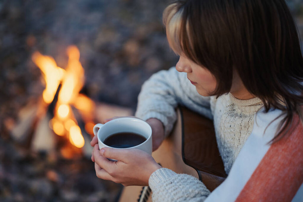 Młoda atrakcyjna dziewczyna gitarzysta z filiżanką gorącej herbaty w pobliżu ogniska. Dziewczyna pije herbatę, gra na gitarze, kemping na świeżym powietrzu w ciepły letni wieczór - Zdjęcie, obraz