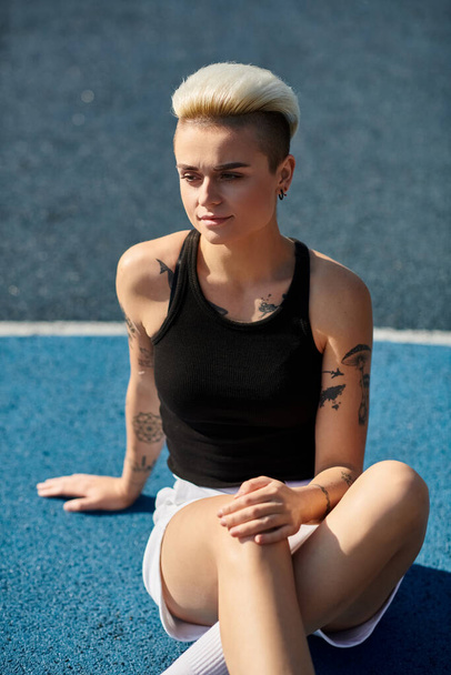 Młoda kobieta z krótkimi włosami i tatuażami siedząca ze skrzyżowanymi nogami na ziemi w spokojnym otoczeniu na świeżym powietrzu. - Zdjęcie, obraz