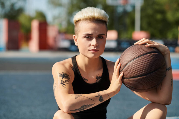Una joven con el pelo corto y tatuajes sentada en el suelo, sosteniendo una pelota de baloncesto, perdida en el pensamiento. - Foto, Imagen