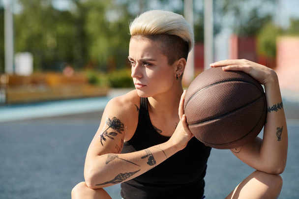 Une jeune femme aux cheveux courts et tatoués s'assoit sur le sol, profondément dans la pensée alors qu'elle tient un ballon de basket dans ses mains. - Photo, image