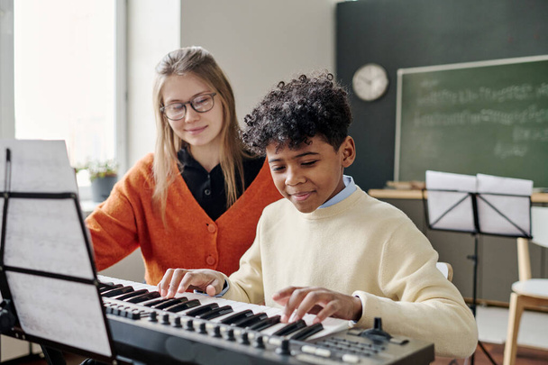 Mediana toma de afroamericano adolescente tocando el piano electrónico en la clase de música, su joven profesor caucásico sentado a su lado - Foto, imagen
