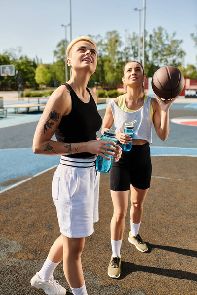 Δύο νεαρές γυναίκες στέκονται με αυτοπεποίθηση σε ένα γήπεδο μπάσκετ, αποπνέοντας δύναμη και αποφασιστικότητα σε σπορ ενδυμασία σε μια ηλιόλουστη μέρα. - Φωτογραφία, εικόνα