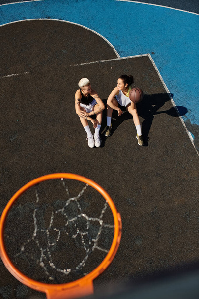 Ένα ζευγάρι αθλητριών φίλων που κάθονται πάνω σε ένα υπαίθριο γήπεδο μπάσκετ μια ηλιόλουστη μέρα. - Φωτογραφία, εικόνα