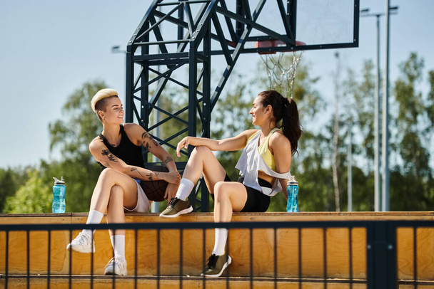İki genç kadın, atletik ve enerjik, güneşli bir açık hava ortamında bir bankta sohbet ediyorlar, bir bağ kurma ve gülme anına dalıyorlar.. - Fotoğraf, Görsel