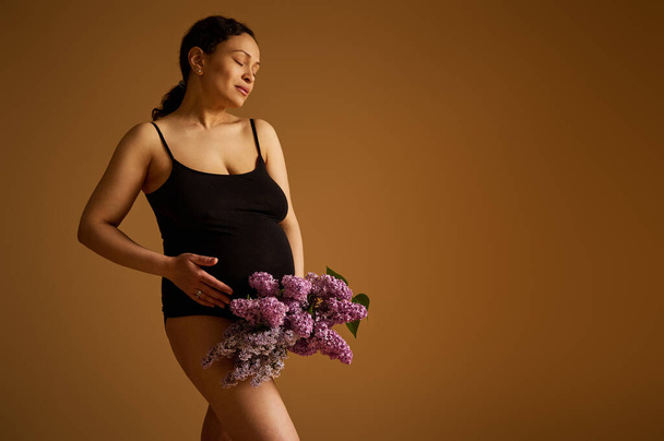 Authentic positive pregnant woman in black bodysuit, smiles while strokes her belly in late pregnancy, last trimester of happy carefree childbearing, posing over isolated beige studio background - Photo, Image