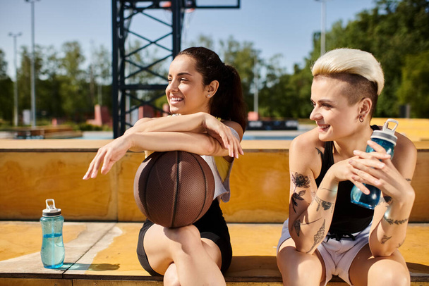 Twee jonge vrouwen met een basketbal zittend op een bankje, bezig met een vriendschappelijk partijtje buitenbasketbal. - Foto, afbeelding