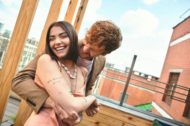 Um homem envolve seus braços em torno de uma mulher em um abraço apertado em um telhado com vista para a cidade, expressando amor e conexão - Foto, Imagem