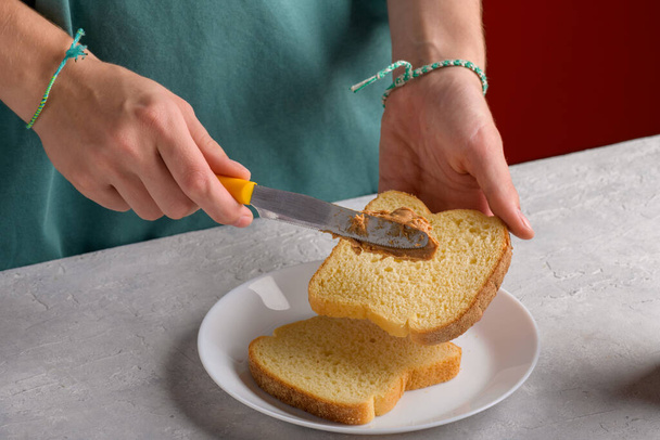 Authentieke vrouwelijke handen die pindakaas verspreiden op een stuk wit brood toast, aan een grijze keukentafel, close-up. Typische snack voedsel, levensstijl, huishoudelijk leven, het bereiden van ontbijt - Foto, afbeelding