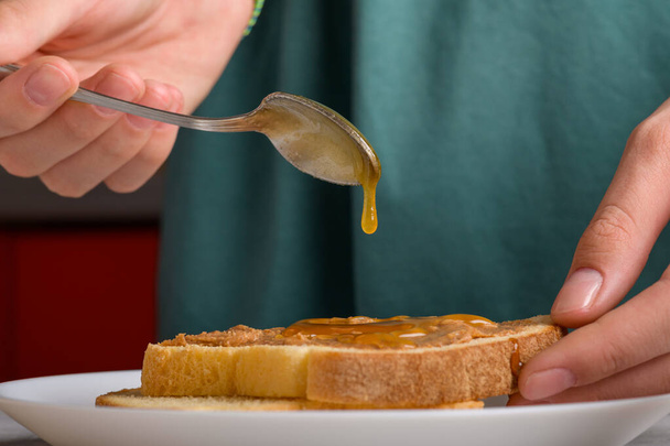 een druppel honing druppelend van een lepel op een boterham met pindakaas. Vrouwen handen die honing met een lepel op een toast zetten met pindakaas om een broodje te maken voor het ontbijt, close-up. Typisch snack voedsel - Foto, afbeelding
