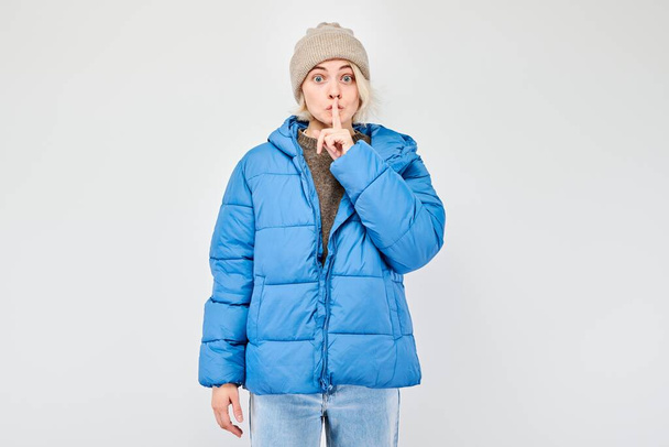 Woman in blue winter jacket gesturing silence with finger on lips, isolated on light background. - Фото, изображение