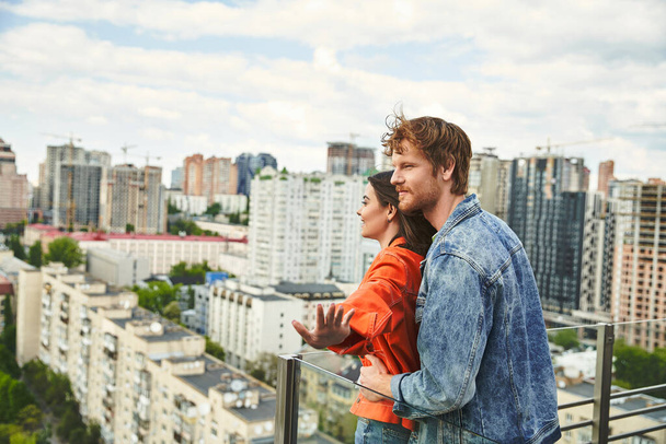 Bir adam ve bir kadın bir balkonda dikilip gökyüzünün altındaki geniş şehre bakıyorlar. - Fotoğraf, Görsel