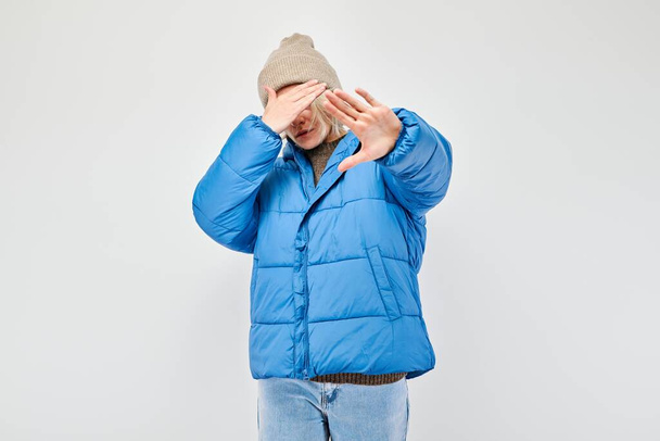 Man in blue jacket and beanie covering face with hands, white background. - Foto, imagen