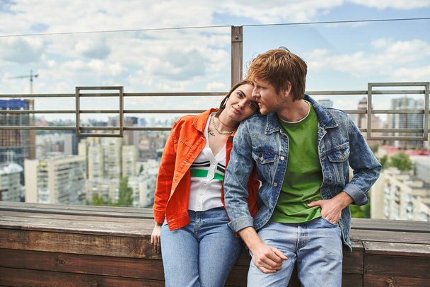 Ένας άντρας και μια γυναίκα κάθονται μαζί στην κορυφή ενός κτιρίου, με θέα το αστικό τοπίο καθώς μοιράζονται μια στιγμή οικειότητας και σύνδεσης. - Φωτογραφία, εικόνα