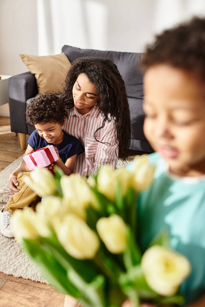 focus on jolly african american mother with son holding gift near her blurred other son with tulips - Photo, Image