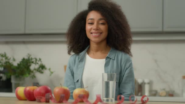 Lächelnd Afroamerikanerin glücklich Frau ethnische Mädchen birassische Dame weibliches Lächeln in die Kamera zu Hause Küche mit Früchten Äpfel Glas Wasser Maßband am Tisch Gewichtskontrolle Ernährung gesunde Ernährung - Filmmaterial, Video