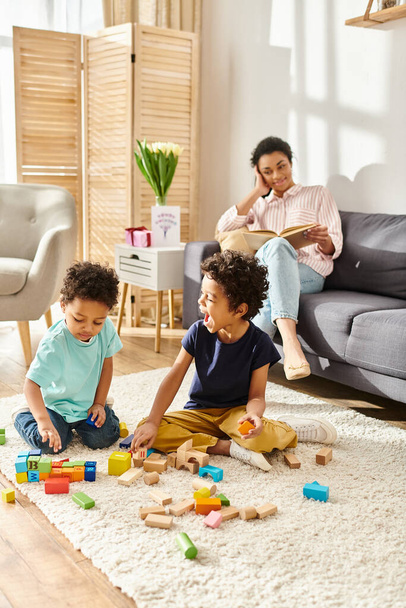 jolly african american woman in homewear reading book and looking at her children play with toys - Foto, imagen