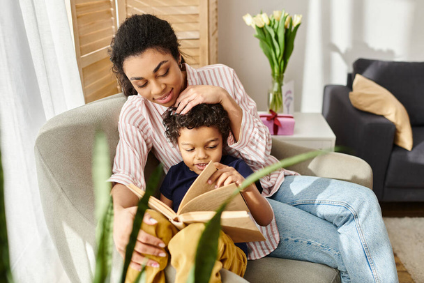 весела приваблива афроамериканка в затишному вбранні читає книгу зі своїм веселим маленьким сином - Фото, зображення