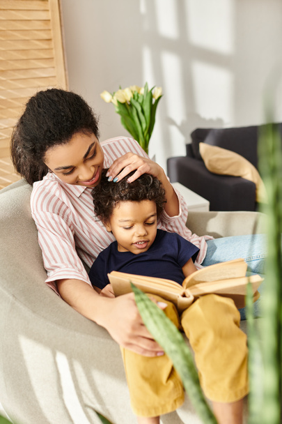joyeuse femme afro-américaine dans le livre de lecture de vêtements de maison avec son petit fils gai à la maison - Photo, image