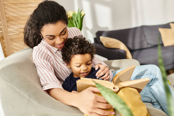 alegre amorosa afro-americana em roupa de casa abraçando seu filho e lendo livro com ele - Foto, Imagem