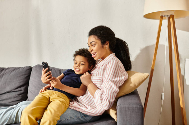 attractive merry african american mother and her adorable son sitting on sofa and looking at phone - Foto, Imagem