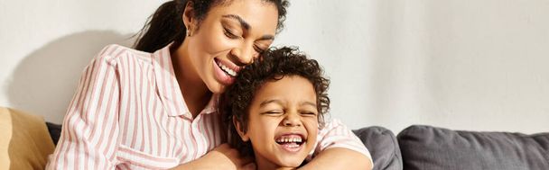 alegre bela mulher afro-americana abraçando seu filho pequeno alegre enquanto na sala de estar, banner - Foto, Imagem