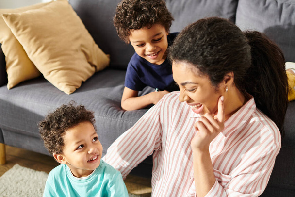 リビングルームにラップトップを持つ彼女の小さな息子の近くに座っているホームウェアで陽気なアフリカ系アメリカ人の母親 - 写真・画像