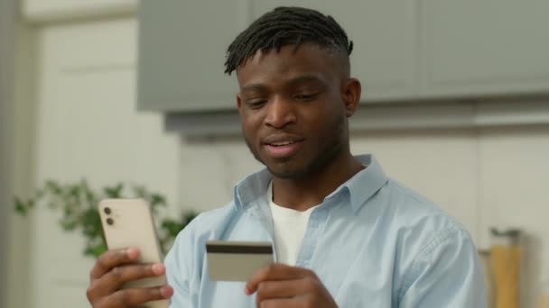 Ευτυχισμένος χαμογελαστός Αφροαμερικανός άνδρας με τηλέφωνο και πιστωτική κάρτα ψώνια σε απευθείας σύνδεση αγορά στο διαδίκτυο αγοράζουν αγαθά μακρινό smartphone τύπος εθνικό αρσενικό αγοραστή πελάτη τραπεζική παραγγελία παράδοση τροφίμων στο σπίτι κουζίνα - Πλάνα, βίντεο