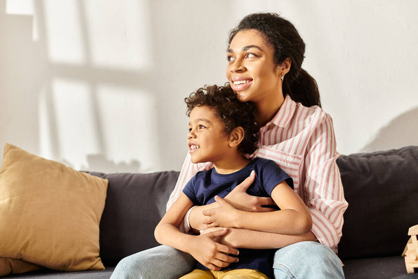 gioiosa madre afroamericana e il suo piccolo figlio che si abbraccia sul divano e distoglie lo sguardo, genitorialità moderna - Foto, immagini