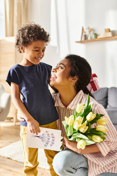 hilpeä afrikkalainen amerikkalainen nainen postikortilla ja kukilla katselee onnellisena poikaansa, äitienpäivä - Valokuva, kuva