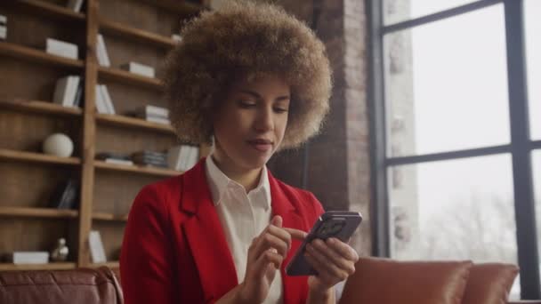 Cheerful businesswoman in a red jacket using her smartphone in an office with a loft aesthetic - Materiał filmowy, wideo