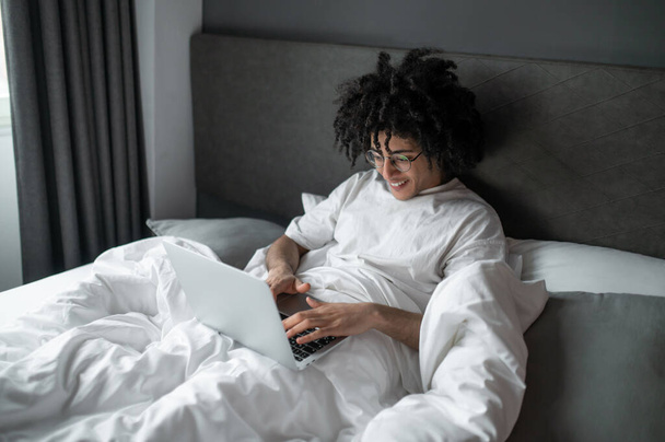 Remote work. Brunette man in white tshirt lying in bed and working on laptop - Foto, Imagem