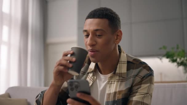 Boldog mosolygós afro-amerikai férfi felhasználó biracial srác etnikai férfi pihentető kanapén otthon mobiltelefon ivás tea kávé ital mosoly chat okostelefon eszköz online vásárlás közösségi média - Felvétel, videó