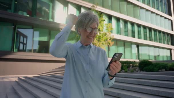 Üst düzey mutlu iş kadını Kafkasyalı kadın açık havada cep telefonu aleti tutuyor. 50 'li yaşlarda heyecanlı bir iş kadını akıllı telefon kazandı. Online zafer dansı başarısını kutluyor. - Video, Çekim