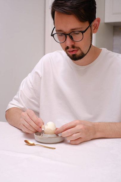 Portrait of a young man peeling an egg at the breakfast table - Photo, Image