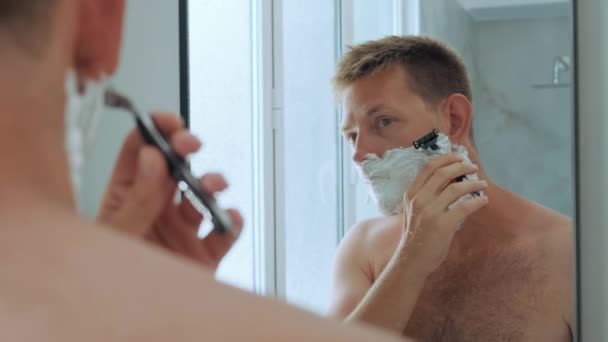 Bianco bell'uomo yong con schiuma da barba sul viso mentre si radeva la barba in piedi in bagno e guardava allo specchio. Giorno mattina procedura di bellezza igienica maschile di routine. - Filmati, video