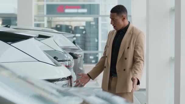 アフリカ系アメリカ人男性男性ビジネスマン ショールームで新しい電気豪華な車モデルのトランクを見てバイヤーを微笑みます 幸せなクライアント ディーラーで自動車販売の自動車を購入することを夢見ています - 映像、動画