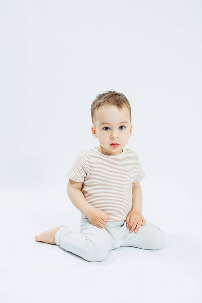 Egy 1 éves kisfiú fehér háttéren ül, és a kamerába néz. Egy fiú pólóban és fehér nadrágban pózol egy könnyű háttéren.. - Fotó, kép