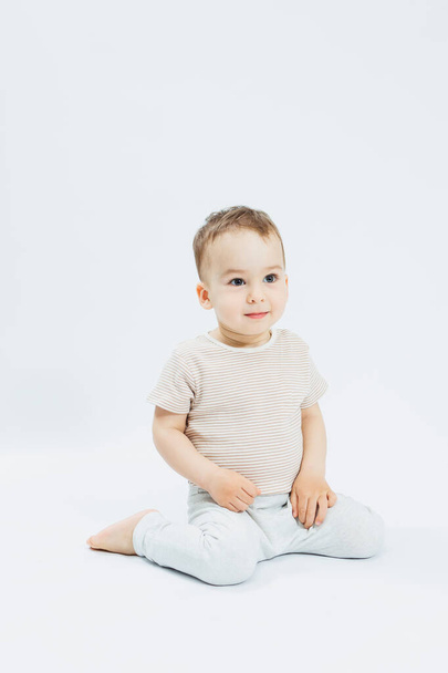 Mały chłopiec w wieku 1 roku siedzi na białym tle i patrzy w kamerę. Chłopiec w T-shircie i białych spodniach pozuje na jasnym tle. - Zdjęcie, obraz