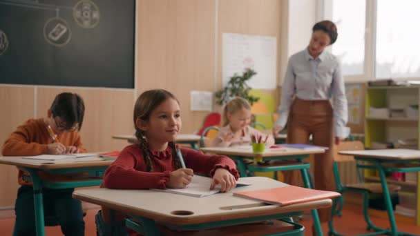 Kaukázusi nő tanár nő tutor séta között osztálytermi asztalok asztal az általános iskolában tanítani a gyerekek segítenek a teszt feladat ellenőrzés gyakorlat notebookok osztály gyerekek írás tanítás tanulás lecke - Felvétel, videó