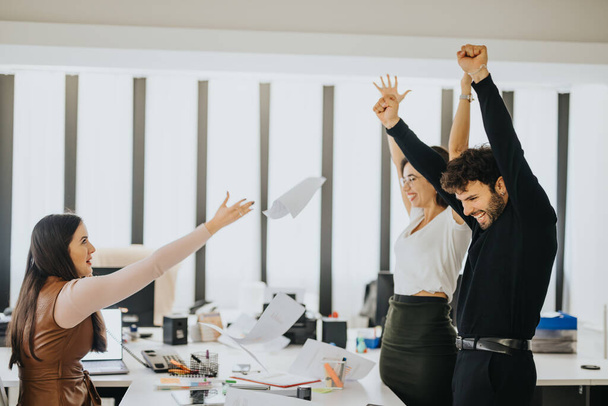 Compañeros de oficina alegres celebrando el éxito con los brazos levantados y papeles volando en un lugar de trabajo moderno. - Foto, imagen
