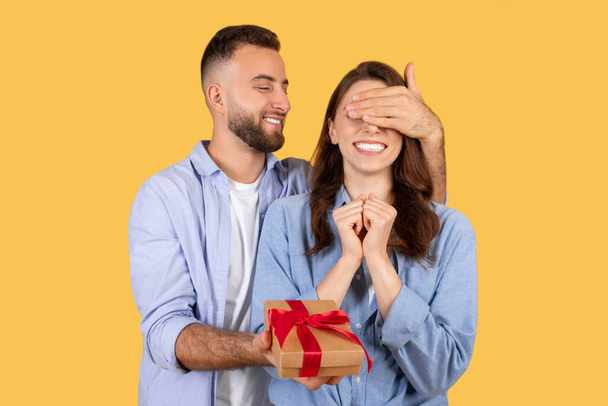 Homme souriant couvrant les yeux des femmes avec sa main, tenant cadeau enveloppé avec ruban rouge, capturant moment de surprise et d'excitation, fond jaune - Photo, image