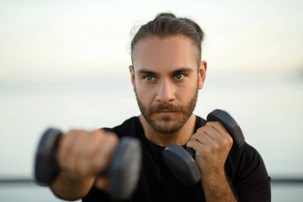Zdeterminowany mężczyzna w fitwear ćwiczenia z hantlami, ucieleśniając aktywny i zdrowy styl życia, zginając bicepsy podczas porannego treningu nad morzem na zewnątrz. Zdjęcie portretowe faceta boksującego z ciężarkami - Zdjęcie, obraz