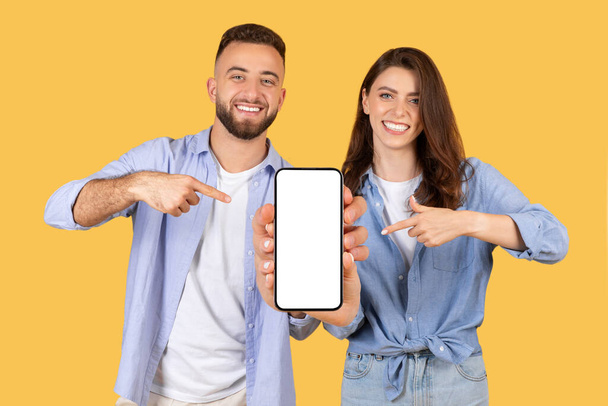 Jeune couple souriant en tenue décontractée et pointant vers l'écran vierge du smartphone, suggérant une application ou un site Web sur fond jaune ensoleillé, maquette - Photo, image
